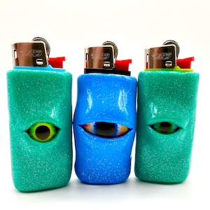 May include: Three custom lighters with eye designs. Two are teal, one is blue, all with glitter. Each lighter has a realistic eye design in the center. The lighters have a silver metal top and a red ignition button.