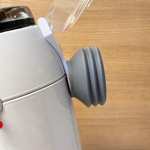 Puede incluir: Primer plano de un electrodoméstico de cocina blanco y gris con una tapa de plástico transparente. Un componente de goma gris y acanalado está fijado al lateral. El electrodoméstico está sobre un fondo de madera marrón claro.