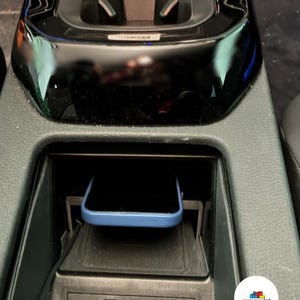 May include: A car's central console featuring a blue smartphone in a charging cradle. A black USB cable is connected to the console. The console is black and grey.