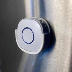 Può includere: Un dispositivo elettronico bianco e grigio con un design circolare blu. Il dispositivo ha una forma arrotondata ed è montato su una superficie metallica. Il design include un pulsante circolare con un contorno blu.
