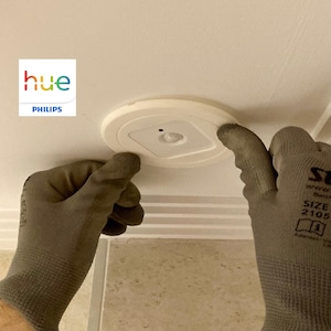 Puede incluir: Un dispositivo de iluminación de sensor de movimiento Philips Hue blanco se está instalando en un techo blanco. El dispositivo de iluminación tiene un sensor cuadrado en el centro y un borde blanco alrededor del borde. La persona que instala el dispositivo de iluminación lleva guantes de trabajo grises.