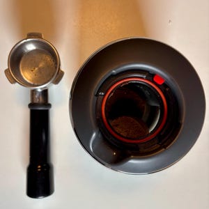 May include: An overhead shot of a disassembled espresso machine. A stainless steel portafilter with a black handle is on the left. On the right is the top of the grinder with a red ring and a small red tab.