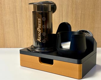 Può includere: Caffettiera AeroPress nera con base nera e arancione. L'AeroPress ha un cilindro trasparente con indicatori di misurazione e il nome del marchio. Sono inclusi anche un portabicchieri nero e un portafiltro.