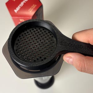 May include: AeroPress XL coffee maker with a black filter and handle. The filter is held over the transparent chamber. A red and white box with the text "AeroPress" is in the background.