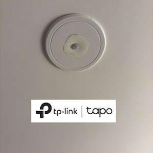 Puede incluir: Un dispositivo inteligente para el hogar TP-Link Tapo montado en el techo, de color blanco. El dispositivo tiene un diseño circular con un sensor más pequeño, blanquecino, de forma cuadrada en el centro. El logotipo de TP-Link Tapo se muestra debajo.