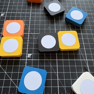 May include: A collection of square-shaped coasters or tiles in various colours including orange, yellow, black, blue, and white. Each piece features a centred white circular insert. The items are arranged on a black cutting mat with grid lines.