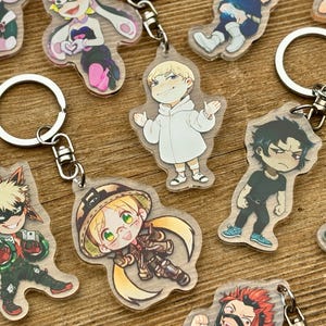 Puede incluir: Una colección de llaveros de personajes de anime con anillos de metal plateado. Los llaveros presentan ilustraciones coloridas de varios personajes en diferentes poses. El fondo es una superficie de madera.