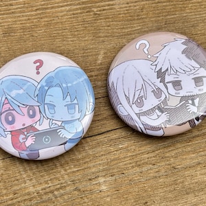 Könnte beinhalten: Zwei runde, farbenfrohe Buttons mit Illustrationen im Anime-Stil. Ein Button zeigt zwei Figuren mit blauen Haaren und einem Fragezeichen. Der andere Button zeigt zwei Figuren in Schwarz-Weiß, ebenfalls mit einem Fragezeichen.