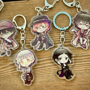 May include: A selection of acrylic keyrings with anime-style character illustrations. Each keyring has a silver-coloured metal chain and a clear, plastic charm with a colourful design. The characters are shown in different poses and outfits.