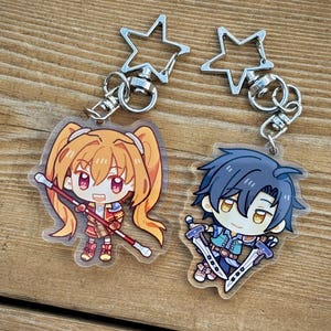 Può includere: Due portachiavi in acrilico in stile anime. Uno presenta un personaggio con i capelli arancioni e un bastone, l'altro con i capelli blu e due spade. Entrambi hanno chiusure a forma di stella e hardware argentato. Lo sfondo è una superficie di legno.