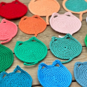 May include: A collection of colorful, handmade crochet cat-shaped coasters. The coasters are in various colors, including red, orange, pink, green, blue, and white. Each coaster has a circular body with ear-shaped cutouts and a tail.