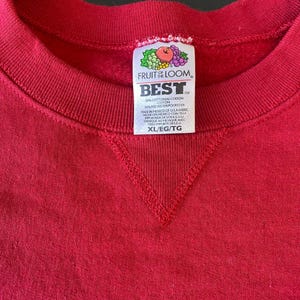 Pode incluir: Uma vista de perto de um moletom vermelho Fruit of the Loom Best. A etiqueta mostra o nome da marca, a composição do tecido e o tamanho XL/EG/TG. O moletom é feito de uma mistura de algodão e poliéster.