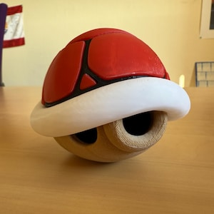 Può includere: Un guscio rosso e bianco con una base in legno. Il guscio è a forma di guscio di tartaruga e ha un contorno nero.