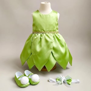 May include: A sleeveless lime green dress with a petal-shaped skirt and a gold-toned waistband. The set includes a pair of matching lime green shoes with white pom-poms and a white headband featuring a decorative bow with sheer fabric accents.