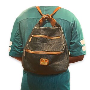 May include: Black leather backpack with three zippered compartments and gold-colored zippers. The backpack has brown leather straps and accents. A gold-colored emblem is visible on the front. The backpack is worn on the back.