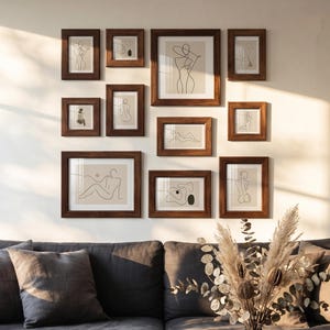 Könnte beinhalten: Eine Sammlung gerahmter Kunstdrucke mit abstrakten Strichzeichnungen in einem Wohnzimmer. Die Rahmen sind dunkelbraun und die Kunstwerke weisen neutrale Farbtöne auf. Ein dunkelgraues Sofa mit Kissen und eine Lampe mit beigem Schirm sind ebenfalls zu sehen.