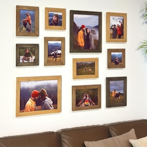 Puede incluir: Una colección de fotografías enmarcadas en varios tamaños y colores, dispuestas en una pared blanca. Los marcos son en tonos marrones y verdes, y las fotos representan escenas al aire libre y personas. Una planta en maceta está en la esquina.