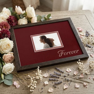 May include: A rectangular dark brown picture frame with a deep red matting featuring the word Forever in white script. The frame contains a photograph of two people leaning against each other. The frame is surrounded by white and dark red roses.