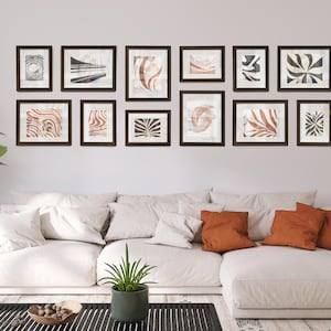 May include: A collection of framed abstract art prints in various sizes and designs, displayed above a white sectional sofa. The artwork features neutral colors and organic shapes, creating a modern and minimalist aesthetic. The frames are dark brown.