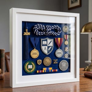 Op de afbeelding: Een witte schaduwdooslijst met een blauwe fluwelen achtergrond en diverse academische memorabilia, waaronder een 2026-kwastje, een gevlochten koord, medailles met de tekst Academic Achievement en Academic Excellence, een schildembleem en spelden.