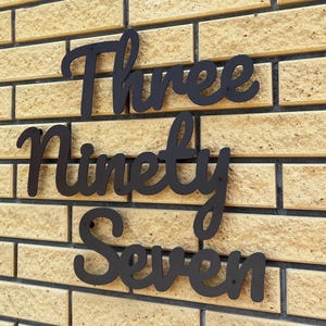 Könnte beinhalten: Schwarze Hausnummern aus Metall, die "Three Ninety Seven" in einer kursiven Schriftart schreiben. Die Zahlen sind an einer hellbraunen Ziegelmauer angebracht.