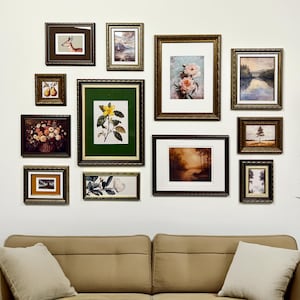 May include: A gallery wall of framed pictures in a living room setting. The frames are dark wood and gold, containing diverse artwork such as landscapes and floral prints. The arrangement is above a beige sofa with cushions. The artwork includes a variety of sizes.
