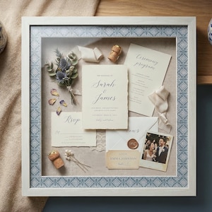 May include: A framed wedding invitation display featuring a bouquet of dried flowers, invitation cards, RSVP card, and a photo. The frame has a white border with a blue and white patterned trim. Corks and ribbons add decorative elements.