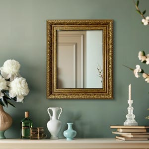 Pode incluir: Um espelho com moldura dourada está pendurado em uma parede verde sálvia, refletindo um vislumbre de um cômodo. Um vaso de flores brancas, um castiçal e vasos decorativos estão em uma prateleira branca sob o espelho. A moldura do espelho tem um design ornamentado.