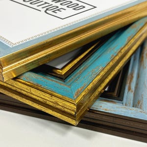 May include: Close-up of a stack of picture frames in gold, distressed turquoise, light blue, and dark brown. The top frame has the text "WOOD BOUTIQUE". The frames are in various styles.