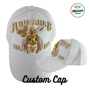 May include: White baseball cap with gold and jewel embellishments. The cap features the name "ABULAHAB" in gold lettering, along with a decorative emblem and the text "205". The words "Custom Cap" are written in black script.