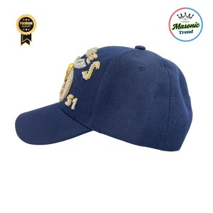 K&ouml;nnte beinhalten: Marineblaue Baseballkappe mit einem gestickten Design auf der Seite. Das Design umfasst goldene und wei&szlig;e Verzierungen. Die Kappe hat einen gebogenen Schirm und einen Knopf oben. Ein Masonic Trend Produkt.