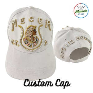 Könnte beinhalten: Weiße, individuelle Baseballkappe mit goldenen und perlenartigen Verzierungen. Auf der Vorderseite steht das Wort "MECCA" über einem Design im ägyptischen Stil mit dem Text "CT. 2". Auf der Rückseite der Kappe steht der Text "HPC 112 HOWARD".