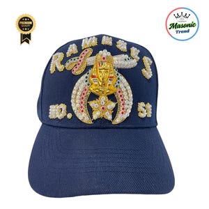 K&ouml;nnte beinhalten: Marineblaue Baseballkappe mit aufwendigen goldenen, roten und wei&szlig;en Verzierungen. Die Kappe tr&auml;gt das Wort "RAMSES" und die Zahlen "10" und "51". Ein goldenes Pharao-Emblem befindet sich mittig auf der Vorderseite.