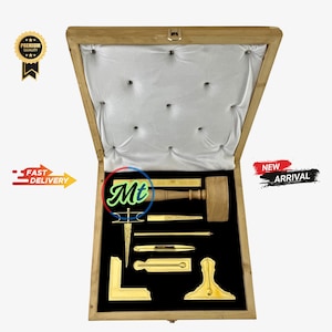 May include: A wooden box with a velvet lining, containing a set of gold-toned tools, including a compass, a protractor, a level, and a mallet. The box is open and the tools are arranged neatly inside.