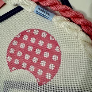 May include: A needlepoint canvas with a pink polka-dot design, accompanied by white and pink embroidery floss. A blue label reads "Natural S503". The canvas is framed with a dark blue border, suggesting a craft project.