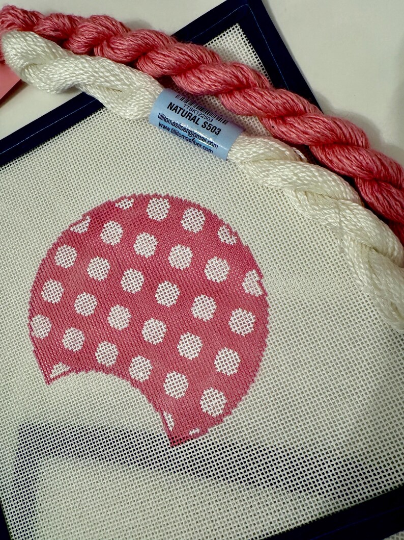 May include: A needlepoint canvas with a pink polka-dot design, accompanied by white and pink embroidery floss. A blue label reads "Natural S503". The canvas is framed with a dark blue border, suggesting a craft project.