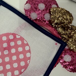 May include: Pink sequined mouse ears with white polka dots and a gold sequined bow. A needlepoint canvas with a pink polka dot design and navy blue border is in the foreground. The canvas has a white background.