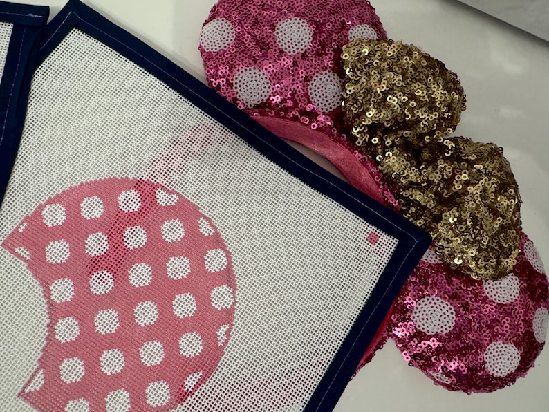 May include: Pink sequined mouse ears with white polka dots and a gold sequined bow. A needlepoint canvas with a pink polka dot design and navy blue border is in the foreground. The canvas has a white background.