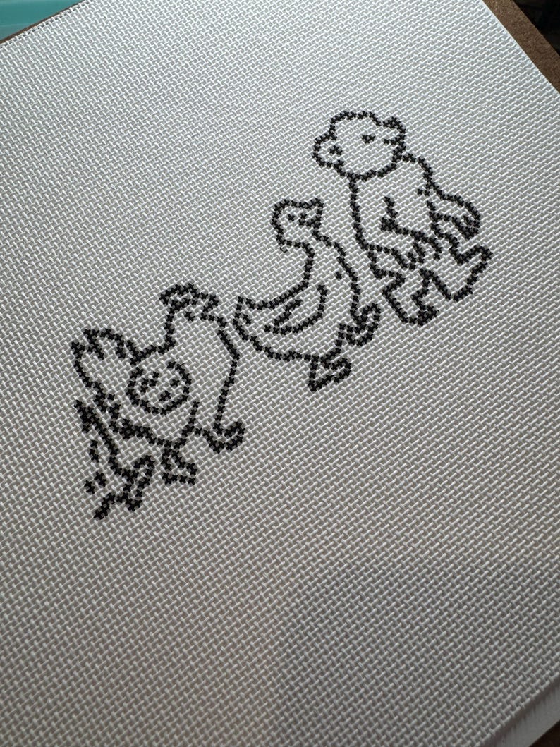 May include: A cross-stitch design featuring a line of three animals: a chicken, a duck, and a bear. The animals are stitched in black thread on a light gray fabric with a woven texture. The design is simple and charming.