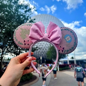 May include: Pink Minnie Mouse ear headband with a large matching bow. The ears have a floral design with pink and green accents. The headband is held up against a blue sky with a large white sphere in the background.