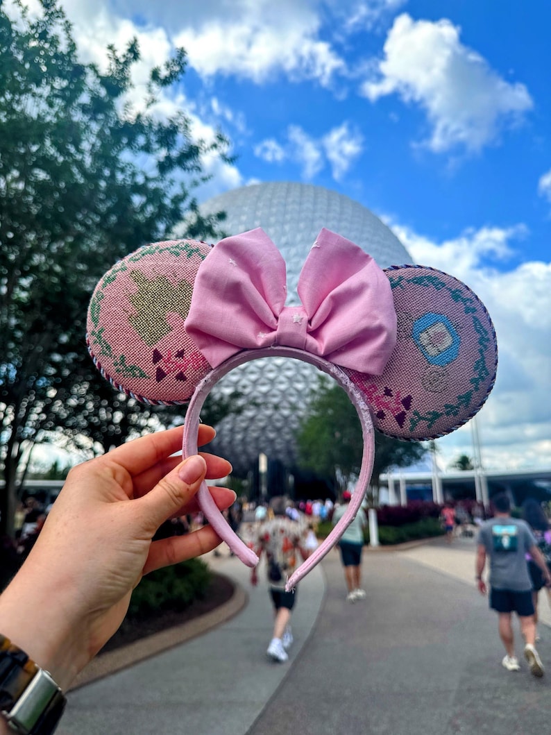 May include: Pink Minnie Mouse ear headband with a large matching bow. The ears have a floral design with pink and green accents. The headband is held up against a blue sky with a large white sphere in the background.