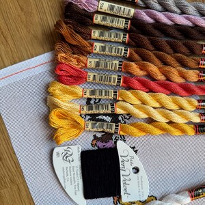 May include: A collection of colorful embroidery floss in shades of gray, brown, orange, red, and yellow, arranged on a wooden surface. A piece of white canvas with a cross-stitch pattern is visible, along with a black and white thread label.