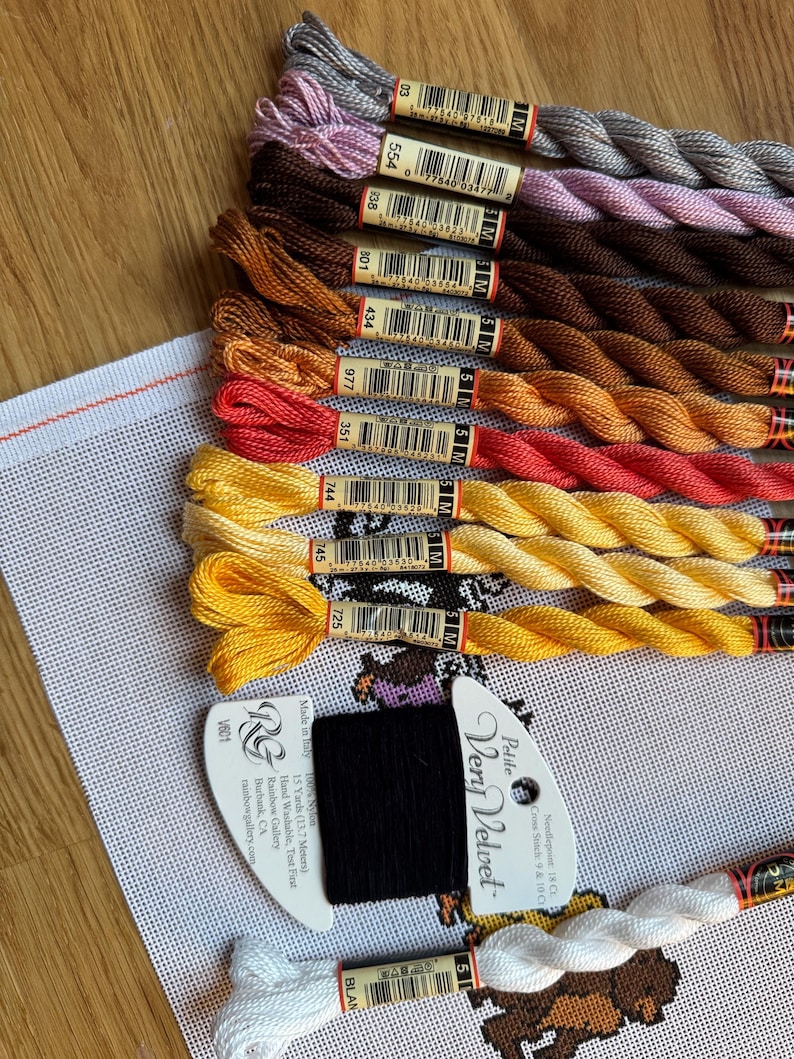 May include: A collection of colorful embroidery floss in shades of gray, brown, orange, red, and yellow, arranged on a wooden surface. A piece of white canvas with a cross-stitch pattern is visible, along with a black and white thread label.