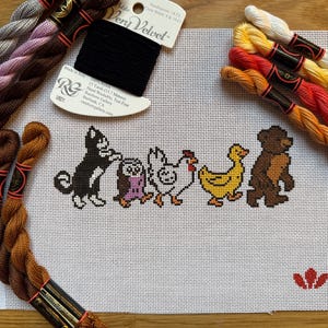 May include: Cross-stitch project featuring a line of cartoon animals: a cat, owl, chicken, duck, and bear. The animals are stitched in black, brown, yellow, and purple on white canvas. Bundles of embroidery floss in various colors surround the canvas.