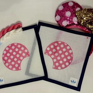 May include: Two square needlepoint canvases with a pink polka-dotted design and navy blue borders. A skein of pink and white embroidery floss and a pair of sequined mouse ears with a gold bow are also in the image.