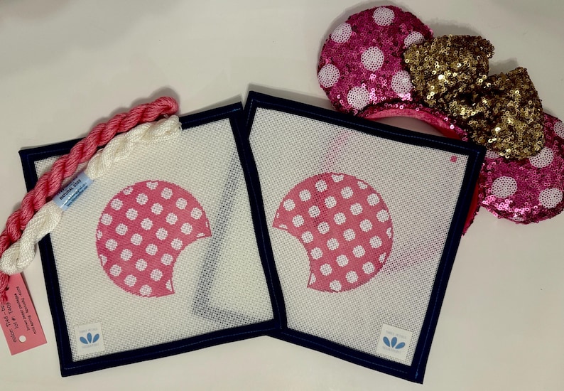 May include: Two square needlepoint canvases with a pink polka-dotted design and navy blue borders. A skein of pink and white embroidery floss and a pair of sequined mouse ears with a gold bow are also in the image.