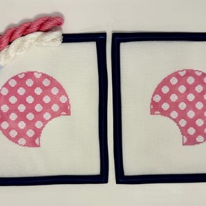 May include: Two square needlepoint canvases with a pink and white polka dot design, framed in navy blue. Two skeins of thread in pink and white are next to the canvases. A tag with text is attached to the thread.
