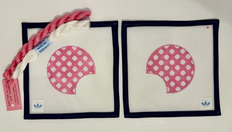 May include: Two square needlepoint canvases with a pink and white polka dot design, framed in navy blue. Two skeins of thread in pink and white are next to the canvases. A tag with text is attached to the thread.