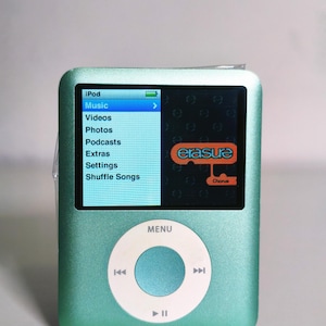 May include: A light green digital music player with a white circular click wheel featuring menu, play, pause, and track skip icons. The screen displays an iPod menu list and an orange Erasure Chorus logo on a dark background with a battery icon.