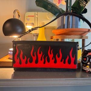 May include: Black leather shoulder bag with a vibrant red flame design. The bag has a long, thin strap and a rectangular shape. A decorative skull mug is next to the bag.